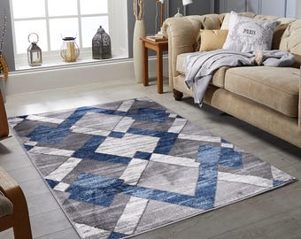 May include: A rectangular area rug with a geometric design. The rug features a pattern of blue, gray, and white shapes. The rug is placed on a wooden floor in a living room setting. The rug's design adds a modern touch to the room's decor.
