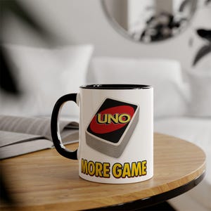 May include: White ceramic mug with a black handle and rim. The mug features a stack of UNO cards with the word "UNO" in red and yellow. Below the cards, the text "MORE GAME" is printed in yellow.