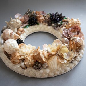 Peut inclure: Une couronne décorative composée de divers coquillages beiges, bruns et crème. Les coquillages sont disposés sur une base blanche texturée, créant un design circulaire. Cette couronne est un élément de décoration intérieure sur le thème côtier.