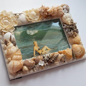 Puede incluir: Marco de fotos cuadrado decorado con varias conchas marinas, coral y flores color crema. El marco rodea una foto de una persona en el océano. El marco es un artículo decorativo con temática de playa.