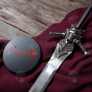 May include: A silver and black sword with a skull-shaped hilt and a matching black display stand. The stand has the text "Devil May Cry" in red. The sword is on a burgundy cloth.