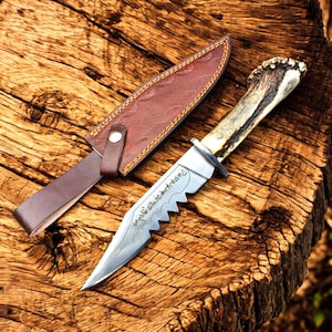 May include: A handcrafted hunting knife with a serrated blade and a deer antler handle. The knife is accompanied by a brown leather sheath. The blade has etched text. The knife and sheath are displayed on a wooden surface.