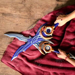 Puede incluir: Dos espadas de fantasía con detalles azules y dorados. Las hojas tienen un diseño curvo con patrones intrincados. Los mangos son de color azul oscuro con detalles dorados. Las espadas se exhiben sobre una tela roja.