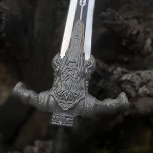 May include: Close-up of a detailed sword with a silver blade and a decorative, gray hilt. The hilt features an ornate design with a bearded face and outstretched arms. The sword's design suggests a fantasy or historical theme.
