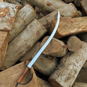 May include: A curved blade sword with a wooden handle and a metal handguard rests on a pile of logs. The blade is silver, and the handle is brown with a patterned grip. The handguard is dark metal. The logs are light brown.