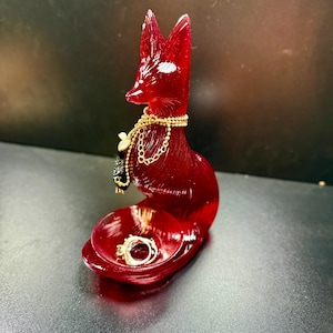 May include: A red fox-shaped ring holder with a gold chain necklace and a small black charm. The holder has a small dish to hold rings. Two rings are in the dish.