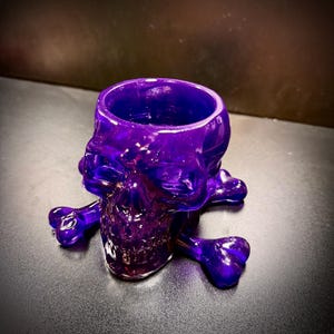 May include: A translucent purple skull-shaped container with a round opening. The skull rests on two crossed bones, also in purple. The container is made of a glossy material and is likely a decorative item or a unique holder.