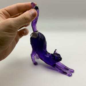 May include: A translucent purple cat figurine in a stretching pose. The cat has a silver ring around its neck and another on its tail. The figurine is held by a hand against a white background.