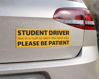 May include: A yellow rectangular bumper sticker with black text reading "STUDENT DRIVER, Not in a rush to learn the hard way. PLEASE BE PATIENT" is affixed to the back of a silver car. The car's taillight is visible.