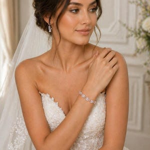 May include: A bride wearing a strapless white wedding dress with intricate lace detailing. She is wearing a delicate bracelet with a floral design and matching earrings. A sheer veil is draped over her hair, and a bouquet of flowers is in the background.