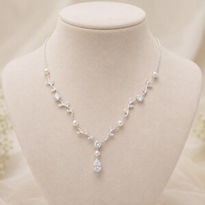 May include: A silver necklace with a delicate floral design, featuring clear crystals and small pearls. The necklace has a teardrop-shaped crystal pendant and is displayed on a mannequin.
