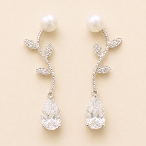May include: A pair of elegant drop earrings. Each earring features a large, pear-shaped, clear gemstone suspended below a curving stem adorned with small, sparkling accents and leaf-shaped details. A large, round, white pearl sits at the top of each earring.