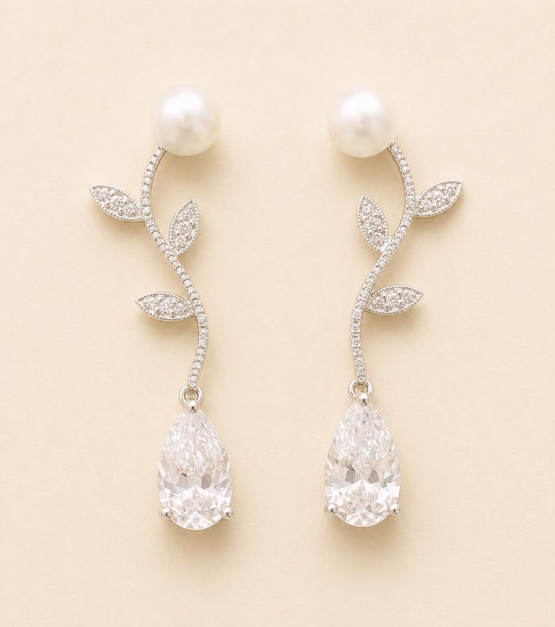 May include: A pair of elegant drop earrings. Each earring features a large, pear-shaped, clear gemstone suspended below a curving stem adorned with small, sparkling accents and leaf-shaped details. A large, round, white pearl sits at the top of each earring.