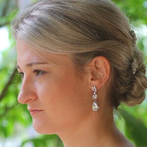 May include: A woman with blonde hair wearing a pair of silver teardrop earrings with clear crystals. The earrings are dangling and have a floral design.