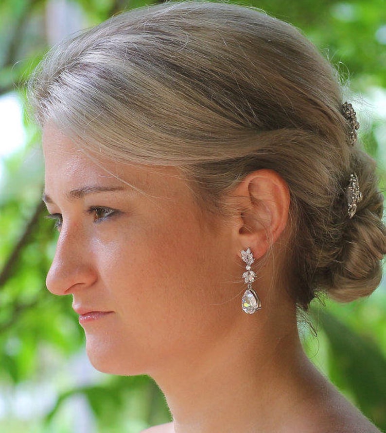 May include: A woman with blonde hair wearing a pair of silver teardrop earrings with clear crystals. The earrings are dangling and have a floral design.