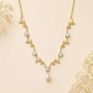May include: A gold-toned necklace featuring a delicate vine design with clear, round gemstones and small leaf accents. A single pear-shaped pearl pendant hangs from the center. The necklace is displayed on a cream-colored background.