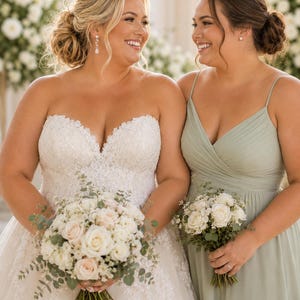 May include: Two smiling women, one in a white strapless wedding gown with a bouquet of white and blush roses, the other in a sage green bridesmaid dress, holding a smaller bouquet. The background features white floral arrangements.
