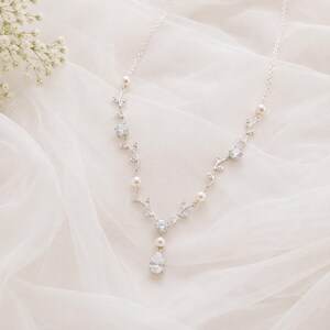 May include: A delicate silver necklace with a floral vine design. The necklace features clear crystals, small pearls, and a pear-shaped crystal pendant. The necklace is displayed on a white, textured fabric, with white flowers in the background.