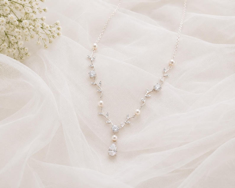 May include: A delicate silver necklace with a floral vine design. The necklace features clear crystals, small pearls, and a pear-shaped crystal pendant. The necklace is displayed on a white, textured fabric, with white flowers in the background.
