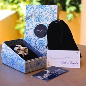 May include: A white gift box with a blue and white snowflake pattern, a black velvet pouch, and a white card with the text "With Thanks" and the company name "Jam Jewels".