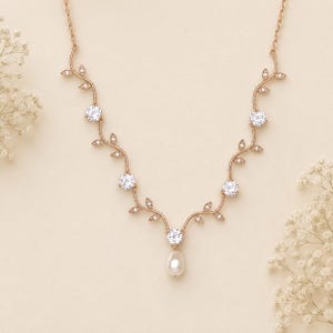 May include: Rose gold necklace with a vine-like design. The necklace features five clear, round gemstones and a single pear-shaped pearl pendant. Delicate leaf accents and small crystals adorn the vine.