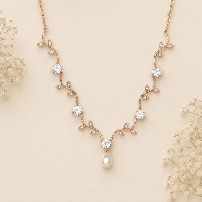 May include: Rose gold necklace with a vine-like design. The necklace features five clear, round gemstones and a single pear-shaped pearl pendant. Delicate leaf accents and small crystals adorn the vine.