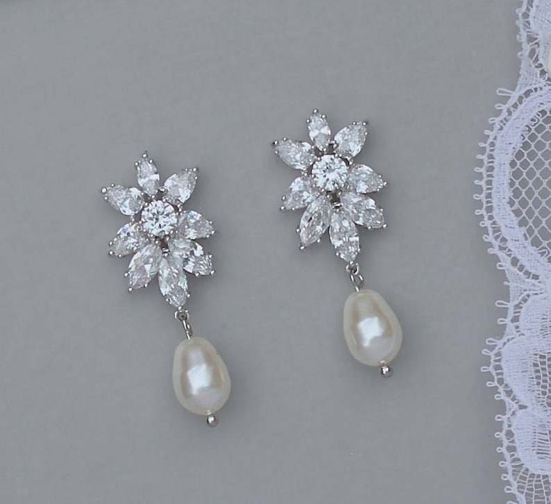 K&ouml;nnte beinhalten: Ein Paar silberne Ohrringe mit einem floralen Design und einem Perlenanh&auml;nger. Die Ohrringe sind aus klaren Zirkonia-Steinen gefertigt.