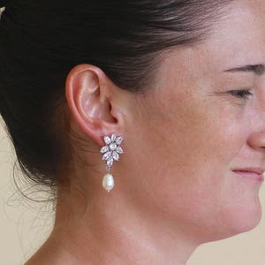 K&ouml;nnte beinhalten: Silberne Ohrringe mit einem Cluster aus klaren Zirkonia-Steinen und einem einzelnen Perlen-Tropfen.