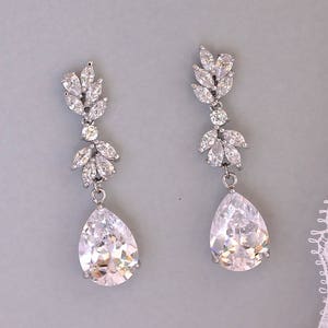 May include: A pair of silver teardrop earrings with clear crystal accents. The earrings feature a cluster of smaller crystals at the top and a larger teardrop-shaped crystal at the bottom.