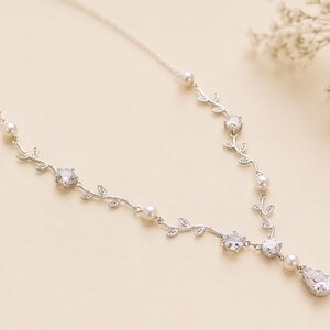 May include: A silver-toned necklace with a delicate vine design. The necklace features clear, sparkling gemstones and small, round pearls. A teardrop-shaped gemstone hangs at the center. The necklace is displayed on a cream-colored surface.