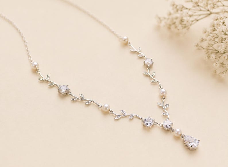 May include: A silver-toned necklace with a delicate vine design. The necklace features clear, sparkling gemstones and small, round pearls. A teardrop-shaped gemstone hangs at the center. The necklace is displayed on a cream-colored surface.