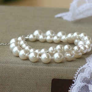 May include: A double strand pearl necklace with a silver clasp. The necklace is made of white pearls and is lying on a brown fabric surface.