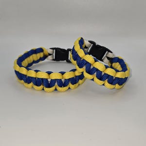May include: Two handmade paracord bracelets. The bracelets are woven in a blue and yellow pattern, with black plastic buckles. The bracelets are displayed on a white surface.