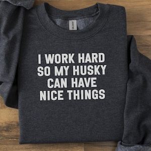 Könnte beinhalten: Dunkelgraues Sweatshirt mit dem weißen Text "I WORK HARD SO MY HUSKY CAN HAVE NICE THINGS". Das gefaltete Sweatshirt liegt auf einer Holzoberfläche. Die Ärmel und der Kragen sind sichtbar. Das Etikett ist am Kragen zu sehen.