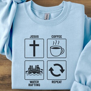 Puede incluir: Sudadera azul claro con un diseño gráfico negro. El diseño incluye una cruz, una taza de café, una escena de rafting y un símbolo de repetición. Las palabras "JESUS", "COFFEE", "WATER RAFTING" y "REPEAT" están impresas.