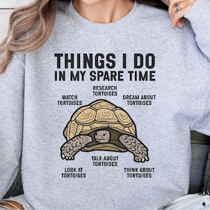 Pode incluir: Uma camisola cinzenta clara de gola redonda com um gráfico de uma tartaruga castanha. O texto diz: THINGS I DO IN MY SPARE TIME, WATCH TORTOISES, RESEARCH TORTOISES, DREAM ABOUT TORTOISES, TALK ABOUT TORTOISES, LOOK AT TORTOISES, THINK ABOUT TORTOISES.