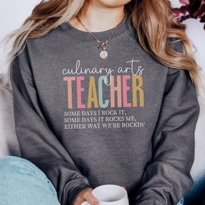 May include: Dark gray sweatshirt with the words "culinary arts TEACHER" in white, pink, yellow, and teal. The text "SOME DAYS I ROCK IT, SOME DAYS IT ROCKS ME, EITHER WAY WE'RE ROCKIN'" is below the word TEACHER.