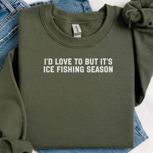 May include: An olive green sweatshirt with the text "I'D LOVE TO BUT IT'S ICE FISHING SEASON" in white. The sweatshirt is folded on top of a pair of blue jeans. The sweatshirt has a crew neck and long sleeves.