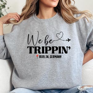 May include: A grey sweatshirt featuring the text "We be TRIPPIN'" in black, with a heart and airplane graphic. Below, the sweatshirt reads "BERLIN, GERMANY". The design is printed on a casual, long-sleeved top.