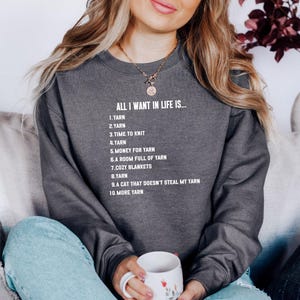 Puede incluir: Sudadera gris oscuro con el texto "ALL I WANT IN LIFE IS..." seguido de una lista que incluye hilo, tiempo para tejer y mantas acogedoras. La persona lleva una taza blanca con un diseño floral.