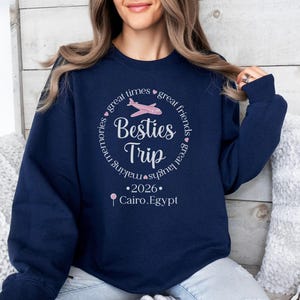 May include: Navy blue sweatshirt with a circular graphic that reads "Besties Trip" with a pink airplane illustration. The text also includes "great times, great friends, making memories, great laughs" and "2026 Cairo, Egypt."