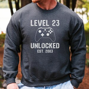 Könnte beinhalten: Dunkelgraues Sweatshirt mit dem Text "LEVEL 23 UNLOCKED EST. 2003" und einer weißen Grafik eines Videospiel-Controllers. Das Sweatshirt hat einen Rundhalsausschnitt.