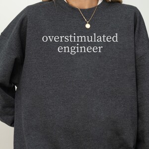 Peut inclure: Sweat-shirt gris foncé avec l'inscription blanche "overstimulated engineer". Ce sweat-shirt à col rond et à manches longues est de couleur unie. Le texte est centré sur le devant du vêtement.