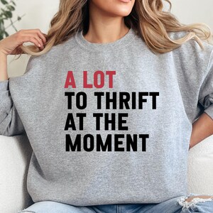 May include: A light gray sweatshirt with the text "A LOT TO THRIFT AT THE MOMENT" in black and red. The sweatshirt is a casual style, suitable for everyday wear. The text is centered on the front.