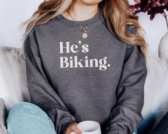 Puede incluir: Sudadera gris oscuro con el texto blanco "He's Biking". La sudadera es de cuello redondo. La persona de la imagen lleva vaqueros azules y un collar dorado. Una taza blanca se sostiene en las manos.