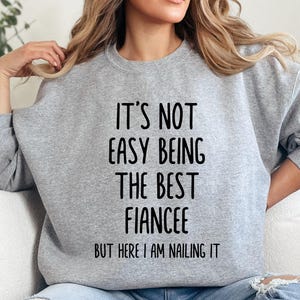 Könnte beinhalten: Hellgraues Sweatshirt mit dem schwarzen Text: "IT'S NOT EASY BEING THE BEST FIANCÉE BUT HERE I AM NAILING IT". Das Sweatshirt hat einen Rundhalsausschnitt.