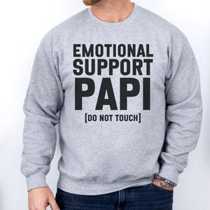May include: A light gray sweatshirt with the text "EMOTIONAL SUPPORT PAPI [DO NOT TOUCH]" in bold black letters. The sweatshirt is a crew neck style and is being worn by a person.