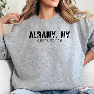 Könnte beinhalten: Hellgraues Sweatshirt mit dem Text "ALBANY, NY" in schwarzen Buchstaben. Darunter die Koordinaten 42.65193° N, 73.75477° W. Das Sweatshirt hat einen lässigen Stil.