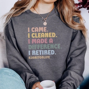 Peut inclure: Sweat-shirt gris foncé avec le texte "I CAME. I CLEANED. I MADE A DIFFERENCE. I RETIRED." en lettres colorées. Le hashtag #JANITORLIFE est en dessous. Le sweat-shirt est de style décontracté.