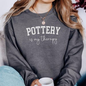 May include: Dark gray sweatshirt with the text "POTTERY is my therapy" in white block and script fonts. The sweatshirt is a crew neck style. A person is wearing the sweatshirt and holding a white mug.